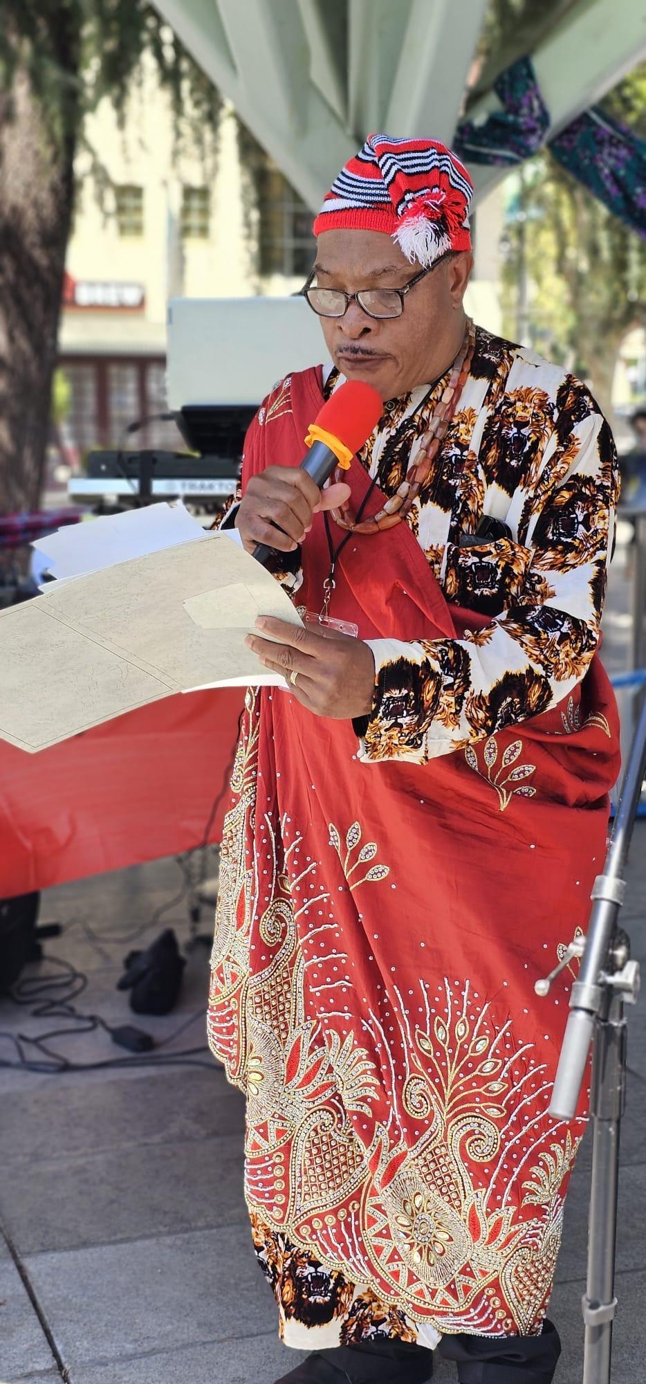 Community member in red traditional attire writing at an outdoor gathering
