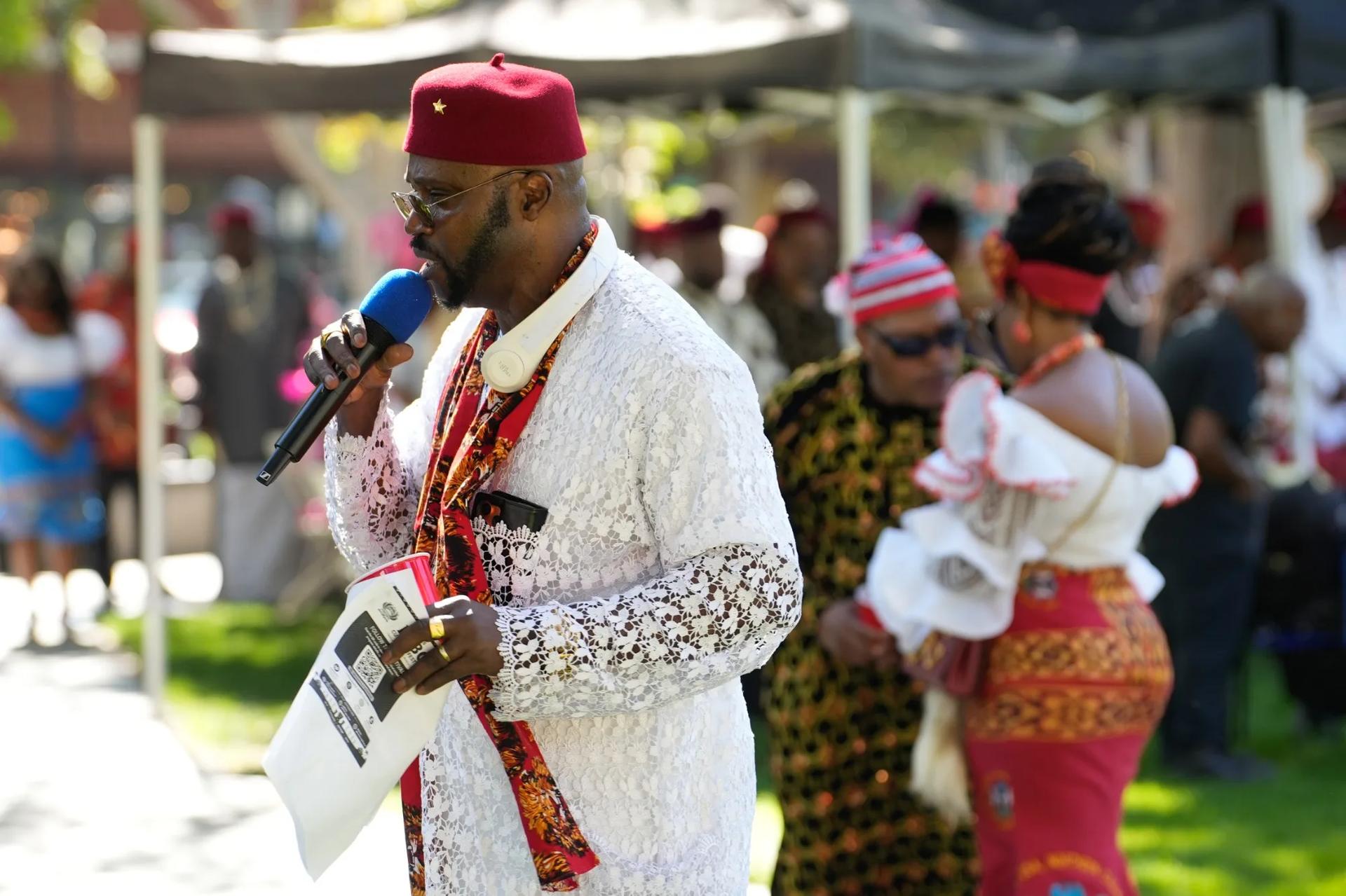 Community elder in white lace attire addressing the festival into a microphone