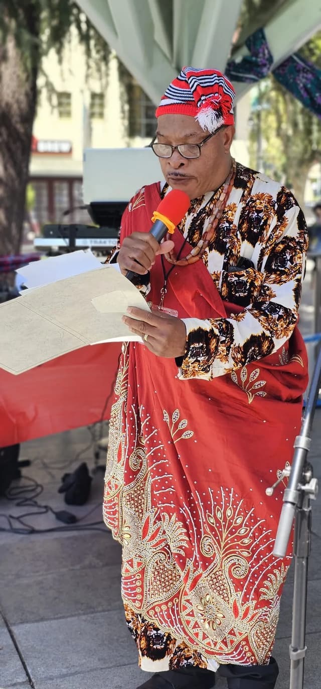 Community member in red traditional attire writing at an outdoor gathering